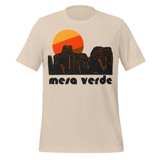 A soft cream colored vintage shirt that says "Mesa Verde", in reference to Colorado's Mesa Verde National Park known for its thousands of archeological sites of the ancestral Pueblo peoples. Includes the image of red rocky outcroppings and southwestern American landscape and a tricolored colorful sun setting behind. 